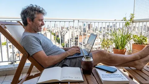 Foto de un hombre sonriente en una reposera de balcón con la laptop, un mate y un cuaderno que dice 'FIRE Argentina'