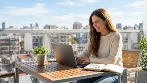 Foto de una mujer sonriente que escribe en la laptop en un balcón con vista a la ciudad, con un cuaderno 'Plan FIRE'